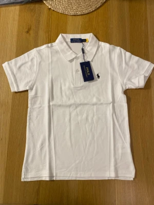 Vit piké t-shirt från Polo Ralph Lauren - Vit piké t-shirt från Polo Ralph Lauren med klassisk krage, två knappar framtill och broderad logga på bröstet. Tillverkad i mjuk bomull och har korta ärmar. Enkel och stilren modell som funkar till det mesta.