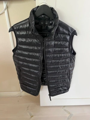 Svart dunväst från Peak Performance - Snygg svart dunväst från Peak Performance med quiltad design och hög krage. Västen har dragkedja framtill och två sidofickor. Perfekt att slänga över en hoodie eller tröja för extra värme. Lätt och smidig att bära med sig.