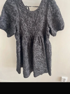 Svart puffärmad klänning med struktur - Säljer en svart klänning med puffärmar och strukturerat tyg. Klänningen har rund halsringning och knytband i ryggen. Materialet är krinklat och ger en cool, trendig look. Perfekt för dig som gillar unika detaljer och volym i ärmarna.