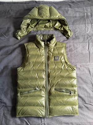 Bvaldi väst - Snygg olivgrön dunväst från Bvaldi med avtagbar huva och två dragkedjeförsedda fickor. Västen har quiltad design, hög krage och ett stilrent logomärke på bröstet. Perfekt för lager-på-lager och håller dig varm under kyliga dagar. De är xs men sitter bra på mig som är 193cm!