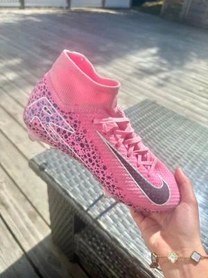 Rosa Nike fotbollskor (mecenat vapo elite)  - Säljer ett par snygga rosa Nike fotbollsskor med högt skaft och svart-vita detaljer. Skorna har ett coolt mönster med svarta prickar och en stor silverfärgad Nike-swoosh på sidan. Perfekta för dig som vill sticka ut på planen och ha bra grepp och passform. Dessa har använts bara 3ggr på en konstgräsplan. Dock har de blivit lite från konstgräset längst fram inte så mycket utan dem är i väldigt bra skick. 