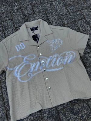 Beige skjorta med tryck  - Kortärmad beige skjorta från  med vit text 'Emotion' och ängel-tryck på framsidan. Skjortan har klassisk krage, knappar framtill och  baksidan är helt beige utan tryck. 