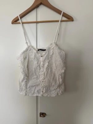 Vit linnetopp med spets från Zara - Söt vit linnetopp från Zara med tunna axelband, knappar framtill och fina spetsdetaljer längs kanterna. Toppen har broderade blommor och en romantisk känsla, perfekt till sommaren. Croppad modell som ger en trendig look.