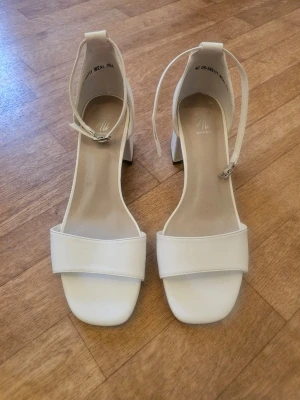 Vita blockklackade pumps heels - Stilrena vita pumps med bred rem över tårna och justerbar ankelrem. Skorna har fyrkantig tå och stabil blockklack. Tillverkade i syntetmaterial med en matt finishoch är oanvända.