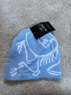 Ljusblå mössa från Arc'teryx - Snygg ljusblå mössa från Arc'teryx med vit logotyp och grafiskt mönster framtill. Tillverkad i mjukt stickat material som sitter skönt på huvudet. Perfekt för kalla dagar när du vill ha både stil och värme.