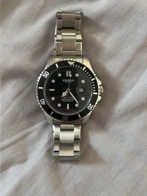 KOMPASS silverfärgad klocka - Stilren klocka från KOMPASS med svart urtavla, datumvisning och tydliga markeringar. Armbandet är i silverfärgad metall och har en klassisk länkdesign. Perfekt accessoar för dig som gillar en tidlös look.