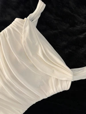 Vit omlottklänning med drapering - Säljer en elegant vit klänning med omlottdetalj och draperad design. Klänningen har breda axelband och är tillverkad i ett mjukt, lätt tyg som ger ett snyggt fall. Perfekt för dig som vill ha en stilren och trendig look.