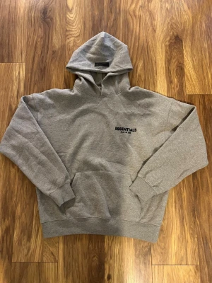 Grå Essentials Fear of God hoodie - Snygg grå hoodie från Fear of God Essentials med svart logga på bröstet och stor text på ryggen. Klassisk passform med huva och känguruficka. Perfekt för en avslappnad streetwear-look. Mjuk bomullsblandning och ribbade muddar.
