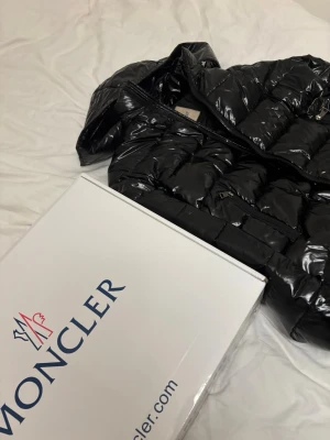 Svart glansig dunjacka från Moncler - Snygg svart dunjacka från Moncler med glansig finish och klassisk quiltad design. Jackan har dragkedja framtill och två praktiska fickor med dragkedja. Perfekt för kalla dagar och ger en riktigt lyxig känsla.