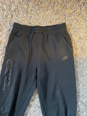 Svarta nike tech fleece byxor  - Svarta mjukisbyxor från Nike med två sidofickor och en extra ficka med dragkedja på benet. Byxorna har smal passform och muddar vid bensluten. Perfekta för en avslappnad och sportig stil.