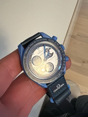 Blå Omega Speedmaster klocka - Snygg Omega Speedmaster klocka med blå boett och svart/blått läderarmband. Klockan har kronograf, tachymeter och tydliga vita detaljer på urtavlan. Perfekt för dig som gillar klassisk design med modern twist.