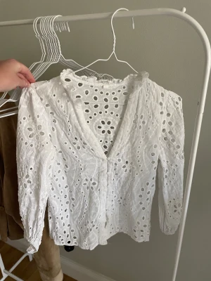 Vit broderad blus med spetsdetaljer - Superfin vit blus med broderade hålmönster och volangkant vid halsen. Blusen har v-ringning, knäppning framtill och långa ärmar med lätt puff. Perfekt att styla med jeans för en fräsch look.