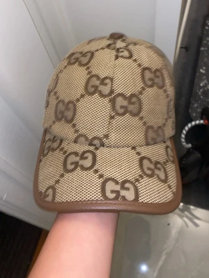 Beige keps från med -mönster - Snygg beige keps med det klassiska monogrammet i brunt över hela kepsen. Kepsen har bruna kanter och justerbar rem baktill. Materialet är canvas med detaljer i läder-look. Perfekt för dig som vill sticka ut med en lyxig accessoar.