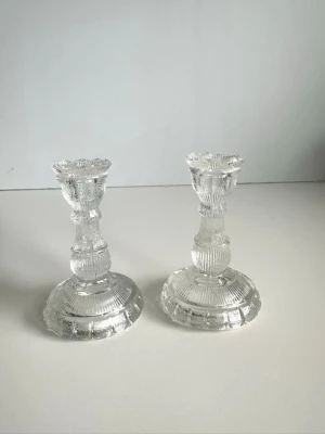 Två ljusstakar i pressglas - Två stilrena ljusstakar i transparent pressglas med dekorativa räfflade mönster och rund fot. Klassisk design som passar i de flesta rum. 10cm höga. Mycket gott skick!