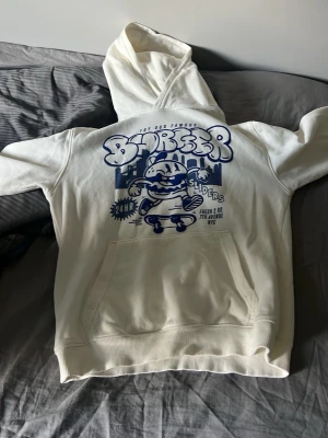 Vit hoodie med blått tryck och grafisk design - Snygg vit hoodie med stor blå grafisk print på framsidan som visar en tecknad figur på skateboard och texten 'Burgers Sliders'. Hoodien har känguruficka och ribbade muddar. Perfekt för dig som gillar streetwear och unika prints.