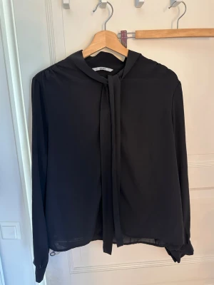Svart blus med knytband i halsen - Stilren svart blus från Only med långa ärmar och knytband i halsen. Blusen har en lös passform och är tillverkad i ett lätt, flowy material som ger en elegant känsla. Perfekt att styla med jeans eller kjol för en trendig look.