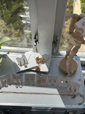 Mix av guldiga örhängen i olika former - Säljer ett set med flera par örhängen i guldig färg. Blandade former som stjärnor, hjärtan, blommor och enkla studs. Några örhängen har små gnistrande stenar och detaljer. Perfekt för dig som gillar att variera din stil och mixa olika smycken.