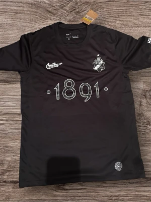 Svart AIK t-shirt från Nike - Svart t-shirt från Nike med AIK:s klubbmärke och texten '1891' på bröstet. Tröjan har rund hals och tryck i vitt. Perfekt för dig som vill visa ditt AIK-stöd på träningen eller på stan.