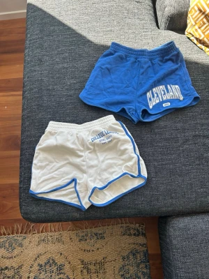College League shorts i blått och vitt - Två par sportiga shorts från H&M, ett par helblå med vit text och ett par vita med blå kant och tryck. Båda har elastisk midja och kort passform, perfekta för träning eller chill. Snygg retrostil med tryck på ena benet.