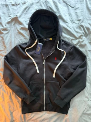 Svart hoodie från Polo Ralph Lauren - Svart hoodie med dragkedja från Polo Ralph Lauren. Klassisk design med vit dragsko i huvan, röd broderad logga på bröstet och känguruficka framtill. Mjuk bomullsblandning som är skön att ha på sig. Perfekt för en avslappnad och stilren look.