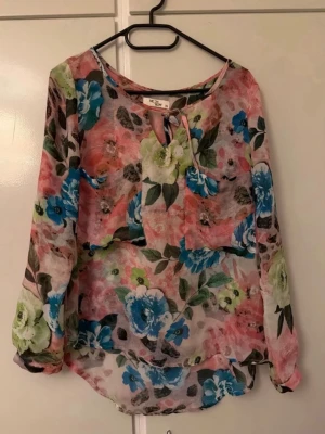 Chiffongblus - Säljer en superfin blommig blus, är transparent med färgglada blommor i blått, grönt och rosa. Den har rund halsringning med knytband och långa, luftiga ärmar. Perfekt till jeans eller kjol för en trendig look.