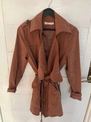 Brun trenchcoat från k.zell - Snygg brun trenchcoat från Kani med avslappnad passform och knytskärp i midjan. Jackan har bred krage, axelklaffar och långa ärmar med uppvikta muddar. Perfekt för dig som vill ha en trendig och stilren look.