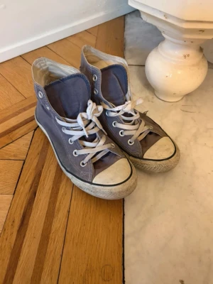 Lila/blå Converse All Star high tops - Klassiska Converse All Star high tops i lila-blå canvas med vita gummisulor och tåhätta. Skorna har vita snören och den ikoniska loggan på sidan. Perfekta för en avslappnad och cool streetstil. Litet hål på sidan i kontakt med sulan (se 3e bild).