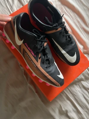 Nike Phantom GT2 fotbollsskor svart/koppar - Snygga Nike Phantom GT2 fotbollsskor i svart och koppar med vita detaljer och rosa dobbar. Skorna har texturerad ovandel för bättre bollkontroll och snörning för optimal passform. Perfekta för dig som vill sticka ut på planen.