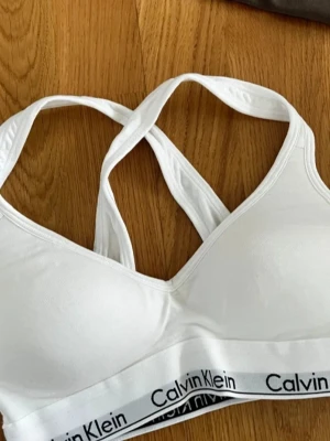 Vit Calvin Klein sport-bh - Vit sport-bh från Calvin Klein. Mjuk och bekväm modell utan bygel i storlek xs. Lätt vaddering som går att ta ut. Den är oanvänd och säljs då den inte kommer till användning.