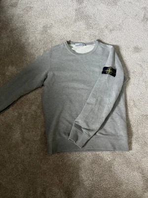 Grå sweatshirt från Stone Island - Säljer en stilren grå sweatshirt från Stone Island med den klassiska svarta patchen med gul och grön logga på vänster ärm. Tröjan har rund halsringning, långa ärmar och ribbade muddar. Perfekt för en clean och avslappnad look.