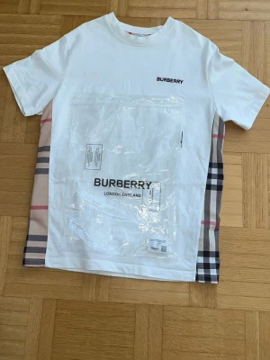 Vit Burberry t-shirt med rutiga sidor helt ny  - Snygg vit t-shirt från Burberry med klassiska rutiga paneler på sidorna och diskret logga på bröstet. T-shirten har rund hals och korta ärmar, perfekt för en trendig och stilren look.