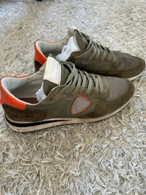 Philippe Model olivgröna sneakers - Snygga sneakers från Philippe Model i olivgrönt mocka och mesh med detaljer i orange och vitt. Skorna har en låg profil, vita innersulor och klassisk snörning. Perfekta för dig som gillar streetstyle och vill ha något unikt på fötterna. Priset är inte hugget i sten
