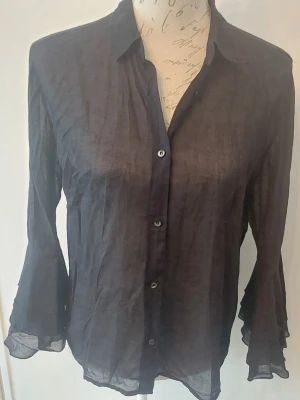 Svart transparent blus med volangärm - Stilren svart blus i transparent material med knappar framtill och klassisk krage. Blusen har långa ärmar med volangdetaljer vid ärmsluten som ger en trendig touch. 