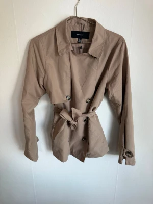 Beige trenchcoat från Vero Moda - Stilren beige trenchcoat från Vero Moda med klassisk krage, knytskärp i midjan och dubbelknäppning framtill. Jackan har långa ärmar och knappar vid ärmsluten. Perfekt för en trendig och tidlös look.
