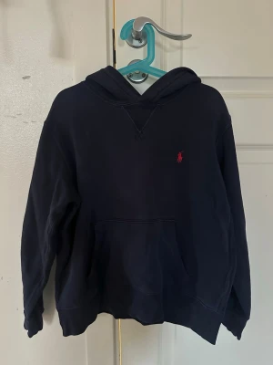 Mörkblå hoodie från Polo Ralph Lauren - Klassisk mörkblå hoodie från Polo Ralph Lauren med röd broderad logga på bröstet. Hoodien har känguruficka framtill och en bekväm huva. Perfekt för en avslappnad stil. Ganska liten i storleken och kan upplevas som en lite bredare modell.
