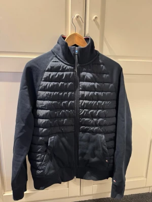 Marinblå quiltad cardigan från Tommy Hilfiger - Snygg marinblå cardiganjacka från Tommy Hilfiger med quiltade paneler framtill och släta ärmar. Jackan har dragkedja, ståkrage och diskreta logodetaljer. Perfekt för lager-på-lager och streetstyle. Materialmix av bomull och syntet.