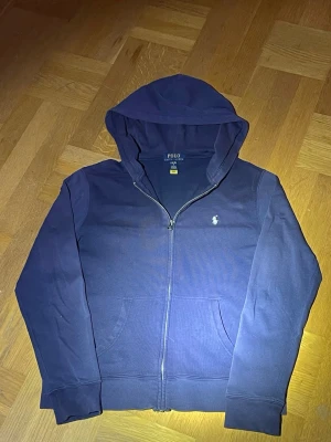 Marinblå hoodie från Polo Ralph Lauren - Snygg marinblå hoodie med dragkedja från Polo Ralph Lauren. Klassisk design med huva, fickor framtill och den ikoniska vita logon broderad på bröstet. Tillverkad i mjuk bomull för skön komfort och stilren look.