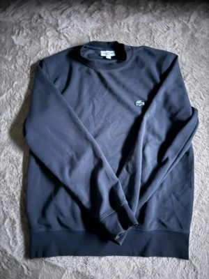 Svart sweatshirt från Lacoste - Klassisk svart sweatshirt från Lacoste med rund halsringning och den ikoniska gröna krokodilloggan broderad på bröstet. Tröjan har långa ärmar och ribbade muddar vid ärmslut och nederkant. Perfekt för en stilren och avslappnad look.