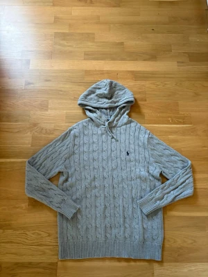 Grå kabelstickad hoodie från Polo Ralph Lauren - Snygg grå kabelstickad hoodie från Polo Ralph Lauren med broderad logga på bröstet. Tröjan har huva med dragsko, långa ärmar och ribbade muddar. Perfekt för en avslappnad och trendig look.