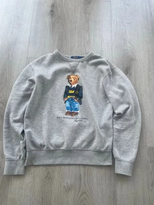 Grå sweatshirt Polo Bear Ralph Lauren - Grå sweatshirt från Ralph Lauren med det klassiska Polo Bear-motivet framtill. Tröjan har rund halsringning, ribbade muddar och är tillverkad i mjukt bomullsmaterial. Perfekt för en avslappnad och trendig stil.