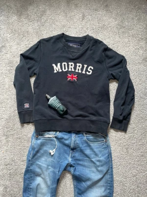 Svart sweatshirt från Morris - Svart sweatshirt från Morris med broderad logga och brittisk flagga på bröstet. Klassisk rund halsringning och långa ärmar. Perfekt för en avslappnad och stilren look.