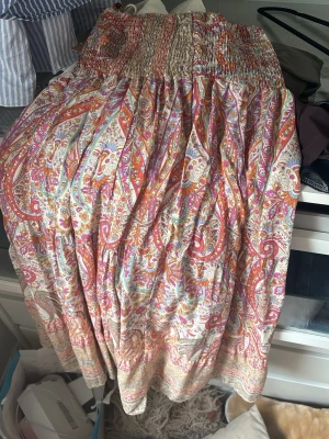 Färgglad paisleykjol från Marta du Château - Säljer en lång kjol från Marta du Château med färgglatt paisleymönster i orange, rosa, lila och turkos. Kjolen har bred resår i midjan och är tillverkad i ett lätt och luftigt tyg, perfekt för varma dagar.