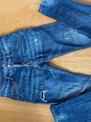 Blå slitna jeans från Dsquared2 - Snygga blå jeans från Dsquared2 med slitningar, små hål och coola detaljer. Jeansen har klassisk femficksmodell, synliga sömmar och en röd logotagg vid gylfen. Perfekta för en trendig streetstyle-look.
