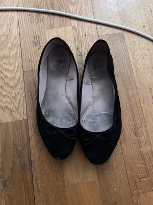 Svarta ballerinaskor med rosett H&M - Klassiska svarta ballerinaskor från H&M med liten rosett framtill. Skorna har rundad tå och är tillverkade i ett mjukt tygmaterial. Perfekta för en enkel och stilren look.