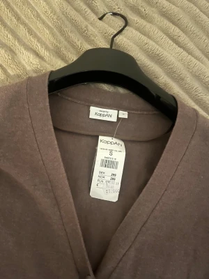 Brun kofta från KappAhl, S - Brun, tunn och mjuk kofta från KappAhl med v-ringning och knäppning framtill. Långärmad modell i ett lätt material som passar perfekt till lager-på-lager. Enkel och stilren design med ribbade muddar vid ärmslut och nederkant.