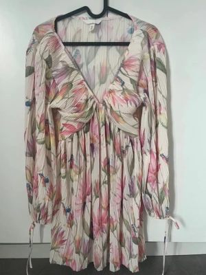 Blommig kortklänning med v-ringning - Superfin kortklänning från H&M med blommigt mönster i rosa, grönt och blått. Klänningen har v-ringning, långa ärmar med knyt vid handlederna och plisserat tyg som ger en luftig känsla. Perfekt för vår och sommar!