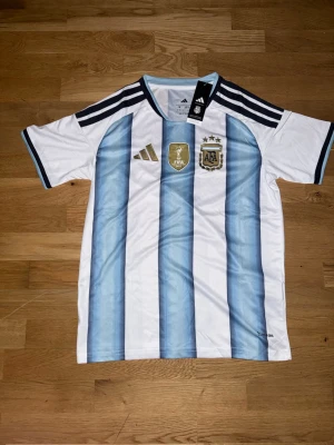 Argentina matchtröja Adidas blå/vit - Säljer en officiell Argentina matchtröja från Adidas med klassiska blå och vita ränder, guldiga FIFA- och AFA-emblem på bröstet samt tre svarta ränder på axlarna. Tröjan är i lätt och ventilerande material, perfekt för fotboll eller träning.