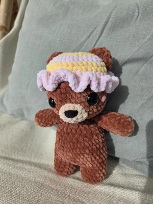 Söt brun nalle med virkad hatt - En mjuk och gosig brun nallebjörn i plysch med stora svarta ögon och beige nos. Nallen har en virkad hatt i pastellfärger, ljusgul och ljuslila, med vågig kant. Perfekt som dekoration eller present till någon som gillar söta saker.