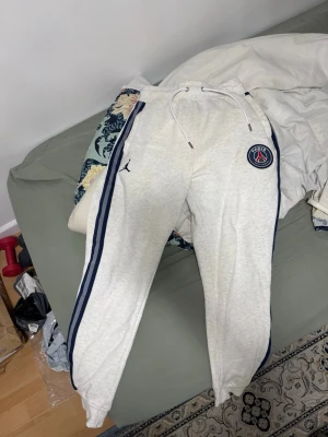 Vita PSG x Jordan mjukisbyxor - Snygga vita mjukisbyxor från Jordan i samarbete med PSG. Byxorna har blå sidoremsor, dragsko i midjan och PSG-logga på ena benet. Tillverkade i mjukt bomullsmaterial, perfekta för en avslappnad stil.