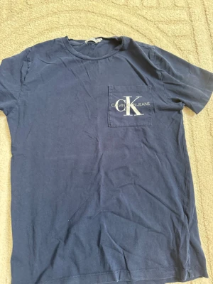 Blå Calvin Klein t-shirt - Mörkblå t-shirt från Calvin Klein med rund halsringning och kort ärm. Bröstficka med vit CK-logga och text. 